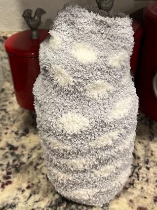 A cozy gray knitted bottle cover with soft, fluffy white spots, placed on a countertop next to red containers.